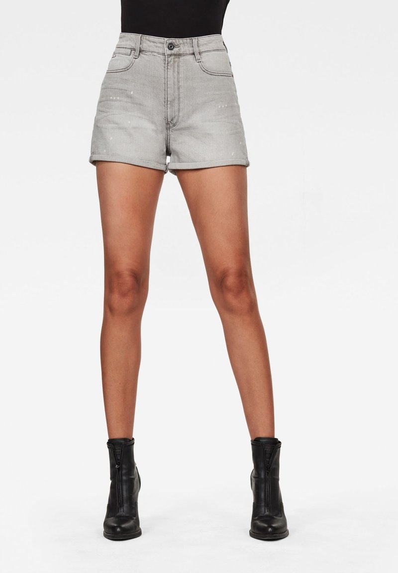 Harmaat farkkushortsit korkealla vyötäröllä, etutaskuilla ja käännetyillä helmillä, yhdistettynä mustiin nilkkasaappaisiin, taustalla yksinkertainen tausta.