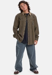 Camisa de cuadros en marrón y negro, con un cuello beige y un corte relajado. Lleva puesta con unos vaqueros azules holgados y zapatillas oscuras.