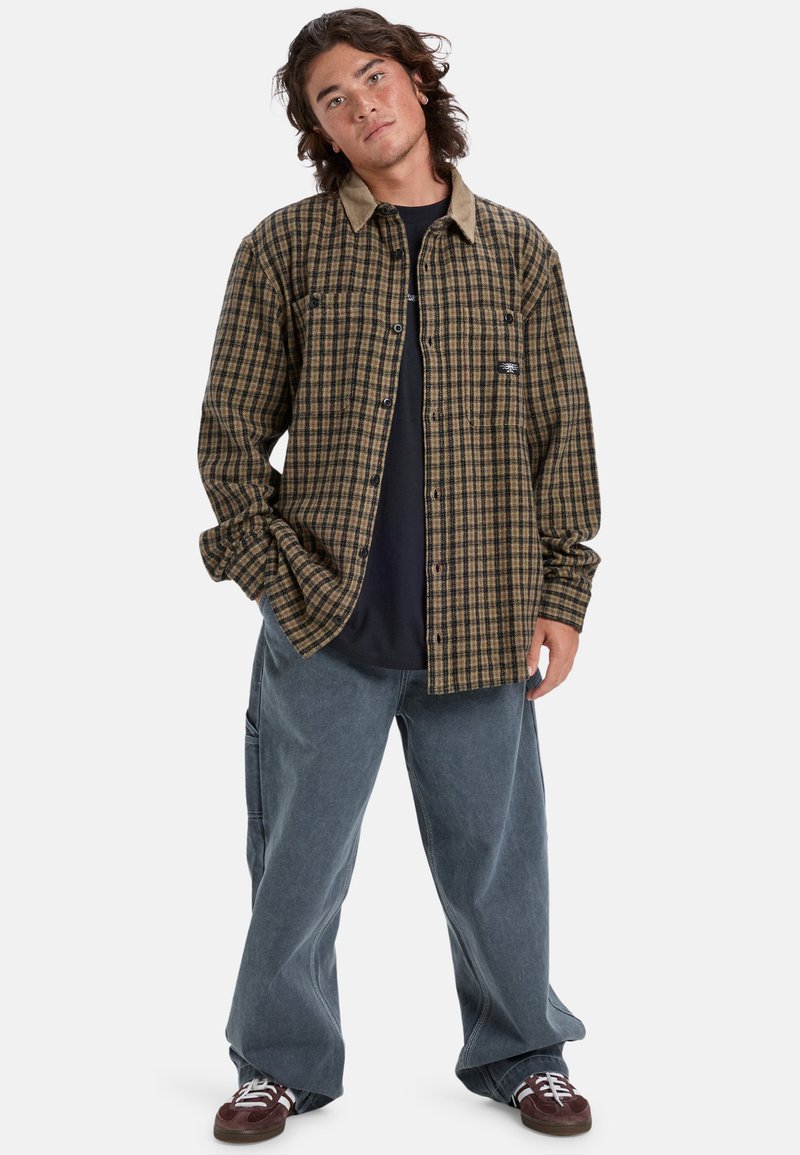 Camisa de cuadros en marrón y negro, con un cuello beige y un corte relajado. Lleva puesta con unos vaqueros azules holgados y zapatillas oscuras.