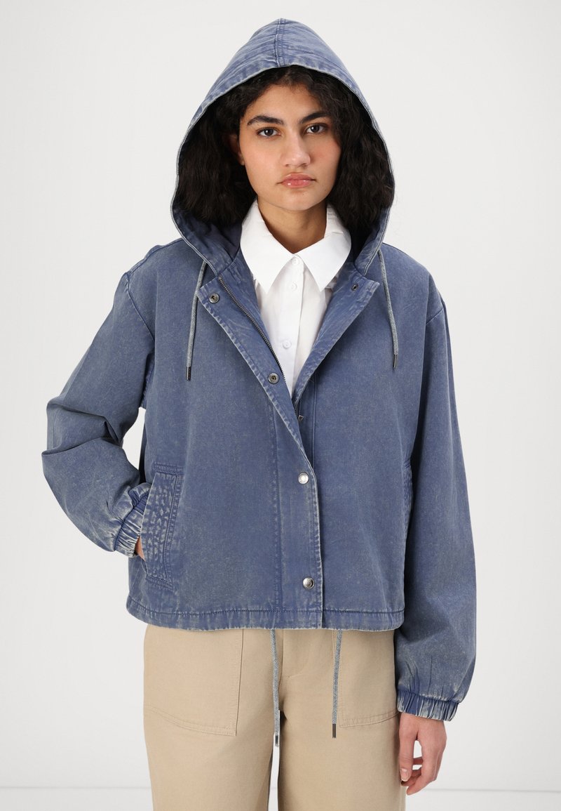 Veste en denim bleu coupée avec capuche, fermeture à cordon, poches avant et poignets élastiques. Présente une texture délavée et des boutons-pression.
