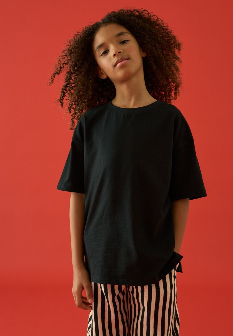 Ragazza con capelli ricci che indossa una maglietta nera larga e pantaloni a righe rosa e nere, in piedi davanti a uno sfondo rosso.