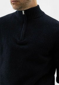 Mann, der einen dunkelblauen, gestrickten Pullover mit einem gerippten hohen Kragen und einem halben Reißverschluss auf einem schlichten, hellen Hintergrund trägt.