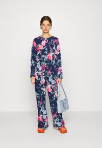 Macaquinho floral com mangas longas, base azul-marinho, flores vibrantes em rosa e verde. Combinado com sandálias de plataforma laranja e uma bolsa azul-clara.