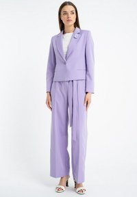 Lawendowy garnitur skrojony na miarę z dopasowaną marynarką i szerokimi spodniami. Posiada broszkę w kształcie kwiatu na klapie oraz gładką fakturę materiału.