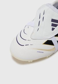 Crampon de football blanc avec un design épuré, une surface texturée, des accents violets et dorés, et une sangle sécurisée sur le cou-de-pied.