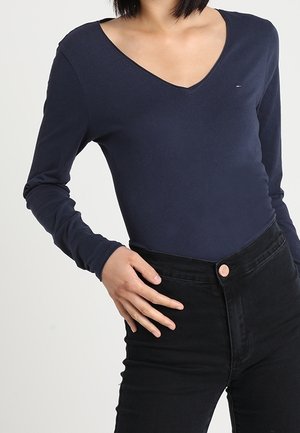 Kobieta ubrana w granatowy, długi rękaw z dekoltem w serek oraz czarne, wysokowalne dżinsy, z ręką opartą na biodrze.