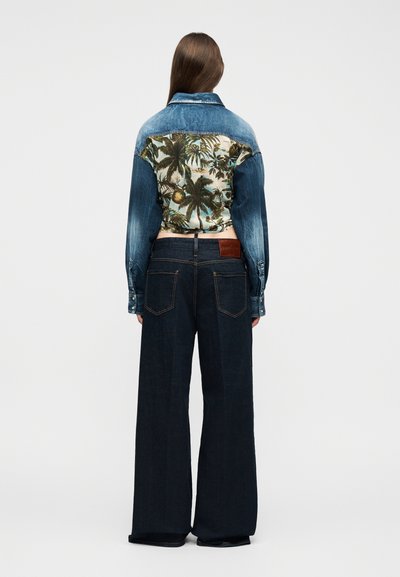 Γυναίκα που στέκεται με την πλάτη προς τα εμπρός, φοράει ένα μπουφάν από τζιν με τροπικό σχέδιο στην πλάτη και φαρδιά μπλε σκούρα τζιν παντελόνια, μπροστά σε απλό φόντο.