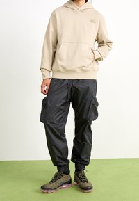 Sudadera beige con un bolsillo canguro y logo, combinada con pantalones cargo negros que tienen bolsillos laterales y puños ajustables. Zapatillas negras y verdes.