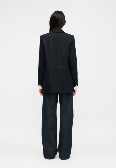 Γυναίκα με μακριά σκούρα μαλλιά φοράει μαύρο σακάκι με διακριτικά κεντήματα και φαρδιά σκούρα παντελόνια, στέκεται μπροστά σε απλό φόντο.