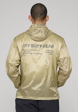 Mand iført en skinnende beige hættetrøje med sort tekst "OFF DUTY GEAR" og mindre skrifter på ryggen, stående mod en ensfarvet baggrund.