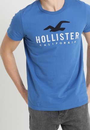 Mand iført en blå Hollister California t-shirt med en sort mågelogo, kombineret med mørkegrå jeans, hænderne hvilende på hoften.
