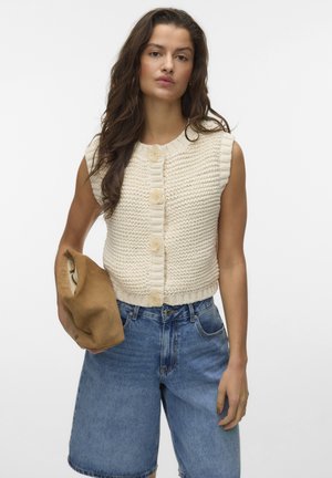 Woman with long dark hair wearing sleeveless cream knit cardigan and loose blue denim shorts holding folded brown bag against white background.