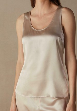 Kobieta ubrana w bezrękawnikowy beżowy satynowy top z zaokrąglonym dekoltem oraz dopasowane satynowe spodnie na jednolitym tle.