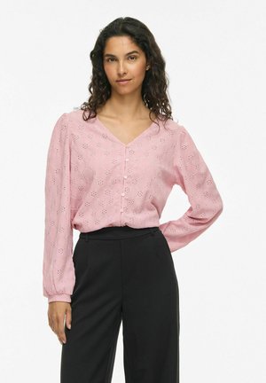 Femme aux cheveux bouclés portant un chemisier rose à manches longues avec des œillets et un pantalon noir taille haute, debout avec une main sur la hanche.