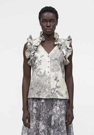 Žena nosiaca bezrukávovú kvetinovú blúzku s volánikmi na ramenách a zladenú kvetinovú sukňu, stojaca na jednoduchom bielom pozadí.