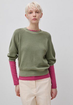 Persona con cabello corto y rubio lleva un suéter de capas verde oliva sobre una camisa de manga larga rosa y pantalones beige claro contra un fondo liso.