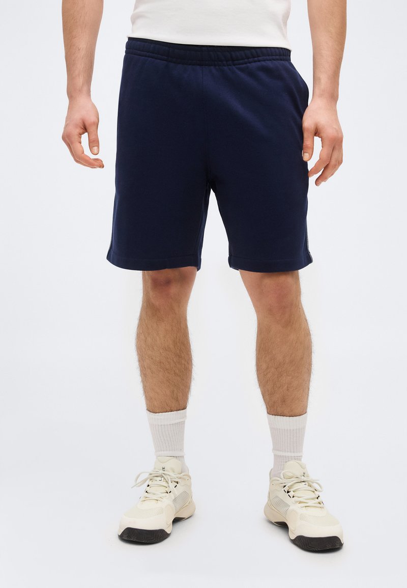 Άνδρας που φοράει σκούρο μπλε σορτς, λευκές κάλτσες μέχρι τον αστράγαλο και λευκά αθλητικά παπούτσια, στέκεται μπροστά σε λευκό φόντο.