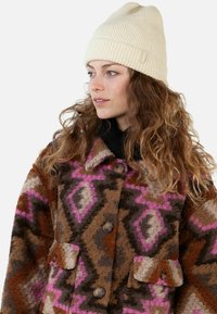 Woman with long curly hair wearing a cream knit beanie and a buttoned patterned coat in brown, pink, and beige tones.