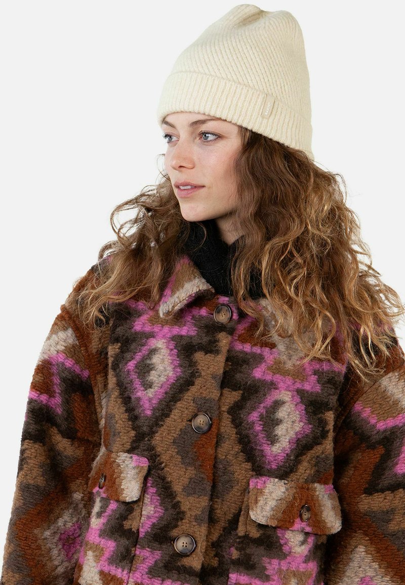 Woman with long curly hair wearing a cream knit beanie and a buttoned patterned coat in brown, pink, and beige tones.