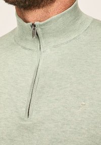 Jasnozielony, dziany sweter z półzamkiem i stójką. Z przodu na piersi małą haftowaną emblemat. Miękka tekstura.