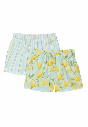 Junge Frau mit dunklem Haar, die ein gelbes Langarmshirt und gestreifte Pastellshorts trägt, steht mit den Händen in den Taschen vor hellem Hintergrund.