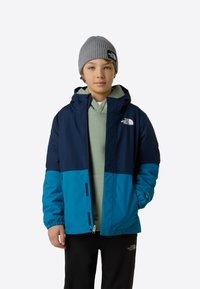 Jachetă în blocuri de culoare albastru și bleumarin, cu glugă, realizată dintr-un material neted, rezistent la apă. Are manșete elastice și detalii cu marcă.