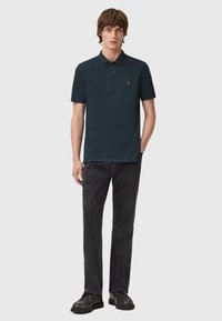 Navy-Polohemd mit Kragen und drei Knöpfen, kombiniert mit schwarzen Jeans und dunklen Schuhen. Moderner Schnitt, glatte Textur, minimales Logo-Detail.