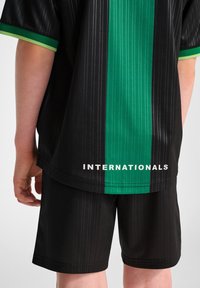 Zwarte sporttrui en shorts met een verticale groene streep en de tekst "INTERNATIONALS" in het wit. Glad, ademend materiaal, gestreept ontwerp.