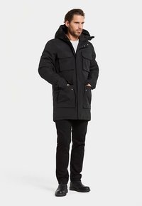 Parka noire avec capuche, dotée de multiples poches avant, d'une fermeture éclair et de poignets ajustables. Portée avec un pantalon et des chaussures noirs.