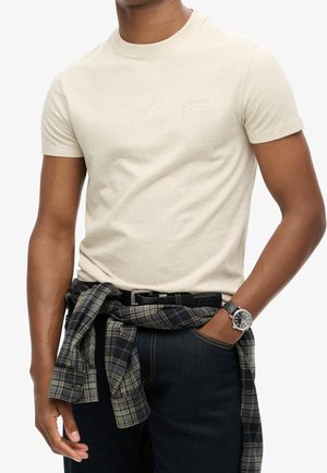 Mand iført lysebeige kortærmet t-shirt, mørke jeans, ur på venstre håndled og en mørk ternet skjorte bundet omkring livet.