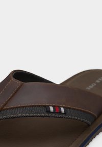 Καφέ λουρί σανδαλιού από δέρμα και ύφασμα με λεπτομέρειες ραφής σε κόκκινο, λευκό και μπλε πάνω σε σκούρο μαλακό πέλμα.