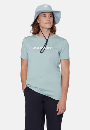 Kvinde iført en lyseblå bredskygget hat med hakestrop og matchende T-shirt med "MAMMUT"-logo, sorte bukser, stående med den ene hånd i lommen.