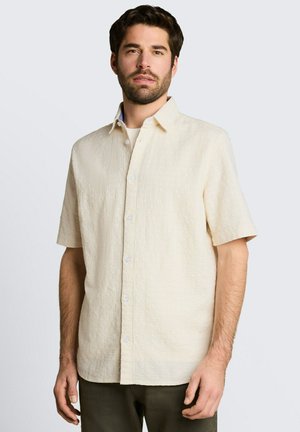 Mężczyzna w beżowej koszuli na krótki rękaw na guziki i ciemnych spodniach, stojący na tle jednolitego jasnoszarego tła.