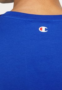 Modrá bavlněná tričko s kulatým výstřihem a malým bílým a červeným logem na zadní straně. Hladký povrch a přiléhavý design.