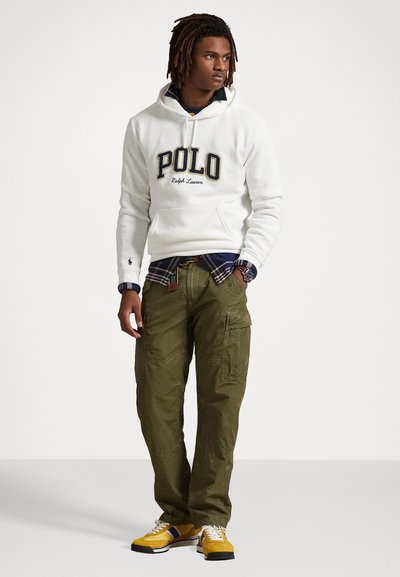 Felpa bianca con logo "POLO Ralph Lauren", camicia a quadri sotto, pantaloni cargo verde oliva e sneakers gialli con dettagli bianchi e blu.