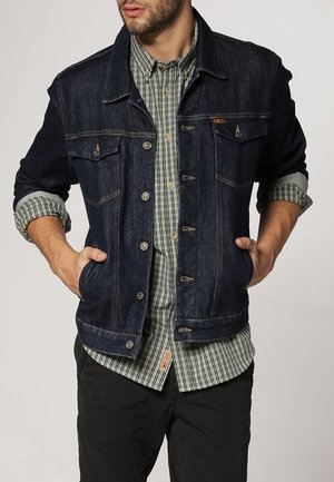 Hombre con chaqueta vaquera azul oscuro sobre camisa a cuadros verde y blanca con mangas arremangadas, con las manos en los bolsillos de la chaqueta.