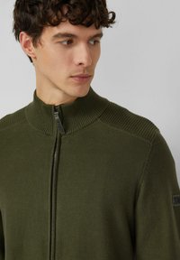 Oliën groene trui met ritssluiting, voorzien van een geribde kraag en schouderaccenten. Gemaakt van zacht materiaal, met een verborgen rits en logodetail.