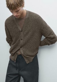 Brown textured cardigan with a V-neck, five buttons, and long sleeves, paired with dark gray pants. Minimal design against a neutral background.