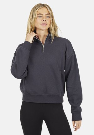 Kobieta w ciemnoszarej bluzie z półzamkiem i długimi rękawami oraz czarnych legginsach, stojąca z jedną ręką przy szyi.