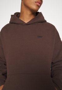 Personne portant un sweat à capuche Levi’s marron avec une poche avant, visible du cou à la taille sur un fond uni.