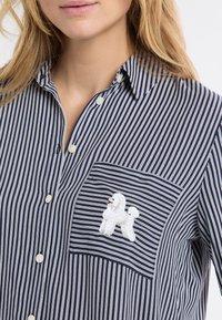 Granatowo-biała pasiasta koszula na guziki z kieszenią na piersi ozdobioną haftowanym wzorem pudla oraz okrągłymi plastikowymi guzikami.