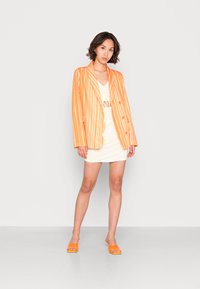 Blazer verticalmente riscado em laranja e branco, combinado com um vestido branco. O modelo usa sandálias de slide laranjas, exibindo um visual contemporâneo.
