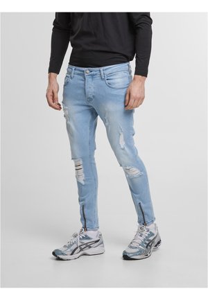 Homme portant un jean skinny déchiré bleu clair avec des zips aux chevilles, des chaussettes blanches, des chaussures de course grises et un t-shirt noir à manches longues, debout sur un sol gris.