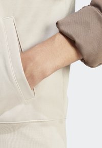 Ruka zasunutá do kapsy béžové mikiny, rukáv hnědého oděvu vyhrnutý, odkrývá předloktí na světlém pozadí.