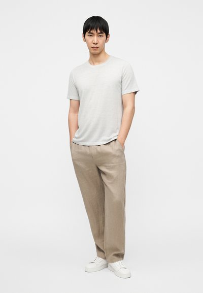 Ανοιχτό γκρι κοντομάνικο μπλουζάκι συνδυασμένο με μπεζ λινά παντελόνια και λευκά αθλητικά παπούτσια. Το μοντέλο στέκεται με τα χέρια στις τσέπες μπροστά σε ένα απλό φόντο.