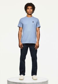 Bărbat cu părul creț, purtând un tricou bleu deschis, blugi închis la culoare și adidași albi, stând pe o platformă rotundă albă.