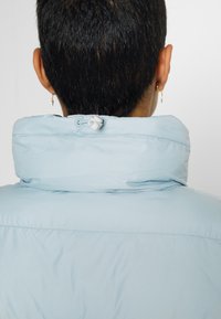 Personne aux cheveux courts et foncés vue de dos, portant une veste bleu clair avec un col haut et un cordon de serrage réglable à l'arrière.