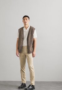 Knit vesta v hnědých a mnohobarevných odstínech s otevřeným předním dílem, nošená přes bílou polokošili a kombinovaná s béžovými kalhotami a černými botami.