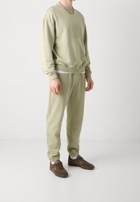 Man draagt een lichtgroene sweatpak met bijpassende joggers en bruine sneakers, staand tegen een effen lichte achtergrond.