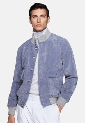 Man wearing a light blue corduroy jacket with gray collar and cuffs over a white turtleneck and white pants, standing against a plain background.